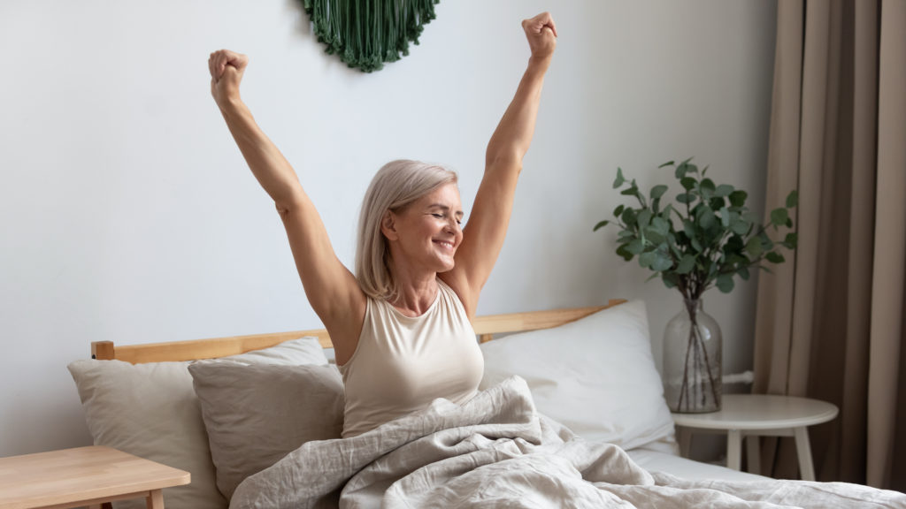 Woman sitting up in bed and stretching with her arms over her head.