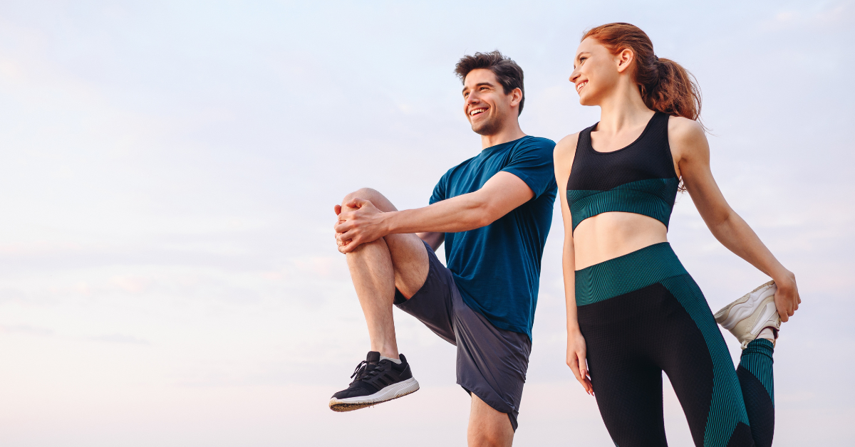 Man and woman in athletic gear smiling while stretching together, appearing to prepare for their workout.