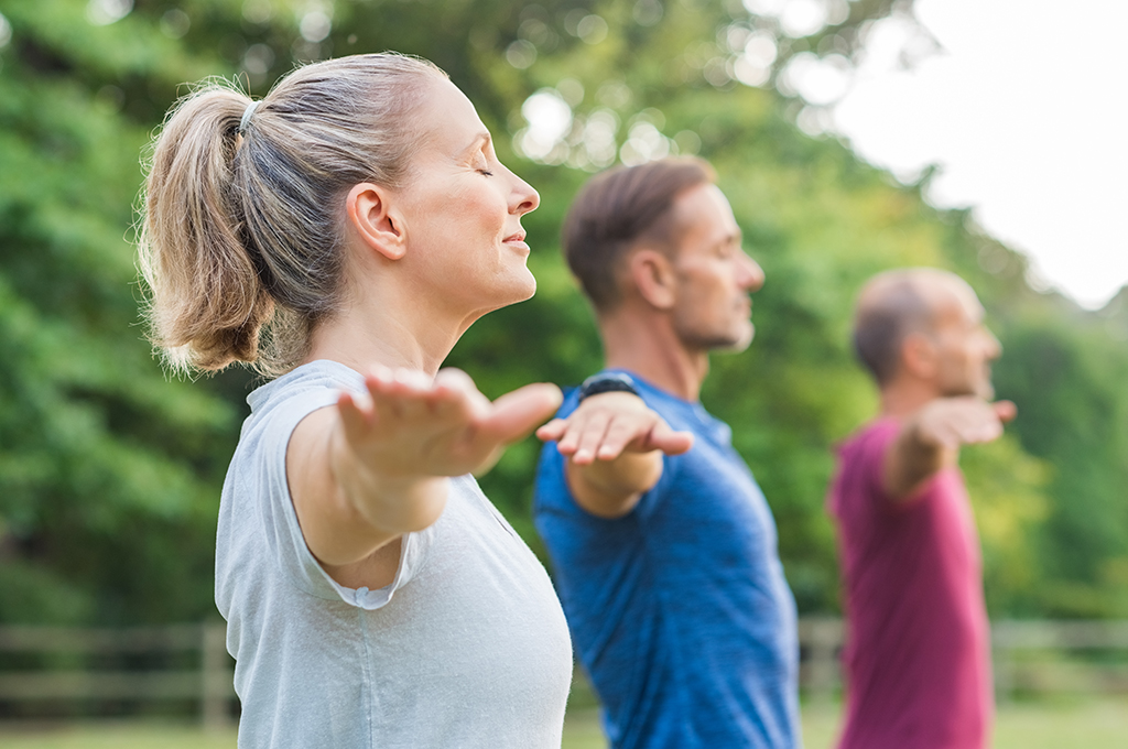 A close-up shot of three people practicing mindfulness or yoga outdoors, standing in a row with arms outstretched, eyes closed, and expressions of calm and relaxation. They are surrounded by greenery, with trees in the background.