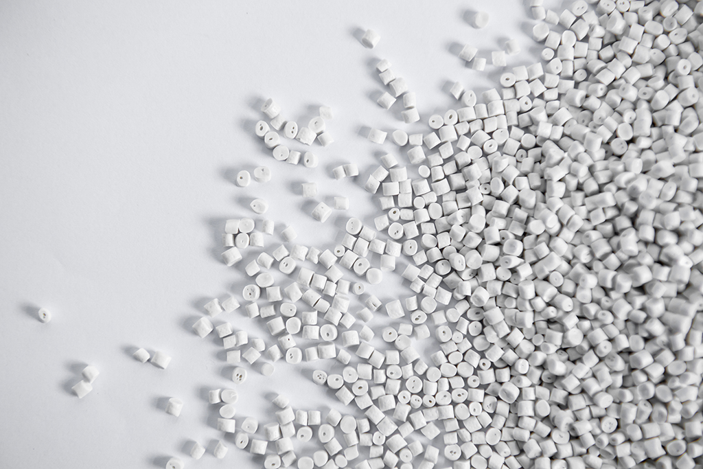 A scattered pile of small, white plastic pellets on a plain white background. The pellets vary slightly in shape, creating a textured pattern against the flat surface.