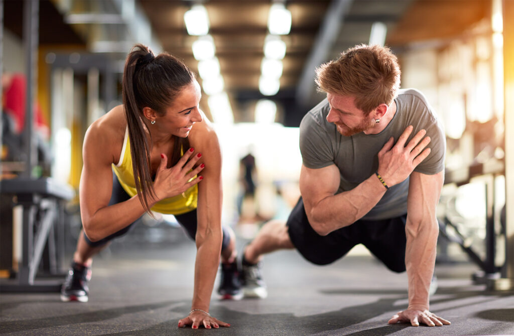 Man and woman in athletic clothing smiling at each other while doing a workout in a gym, appearing to enjoy the exercise together.