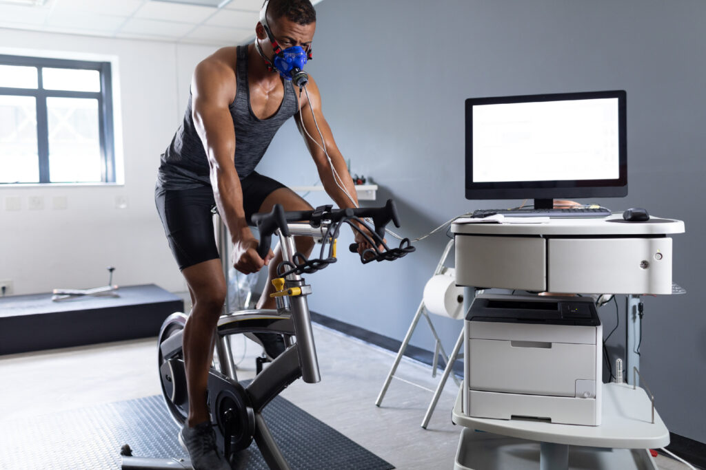 Man wearing a breathing mask and sensors while riding a stationary bike in a lab setting. A computer and testing equipment are visible beside him, indicating a fitness or performance test.