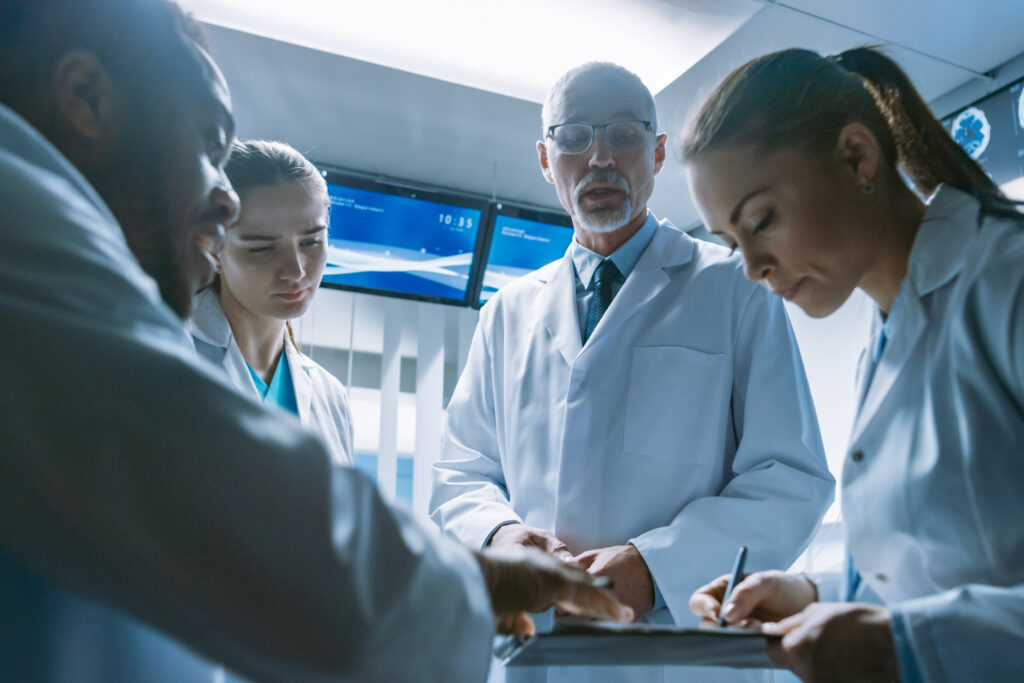 Group of four people—two men and two women—wearing lab coats, gathered around as one person takes notes on a clipboard, appearing to discuss or analyze data in a laboratory setting.