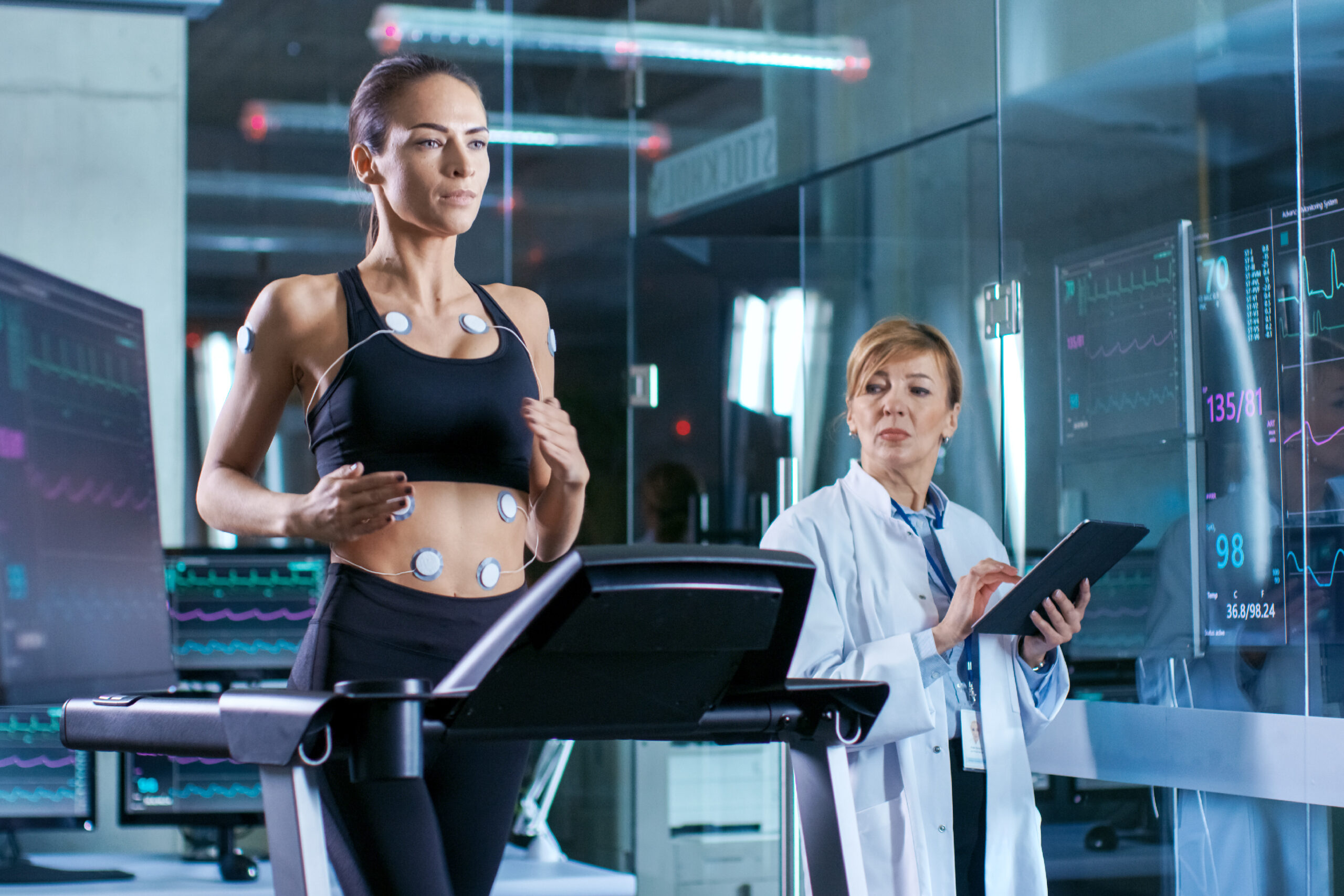 Woman in athletic clothing running on a treadmill with diagnostic sensors attached to her body, while a female technician in a lab coat records data on a clipboard in a clinical or research setting.