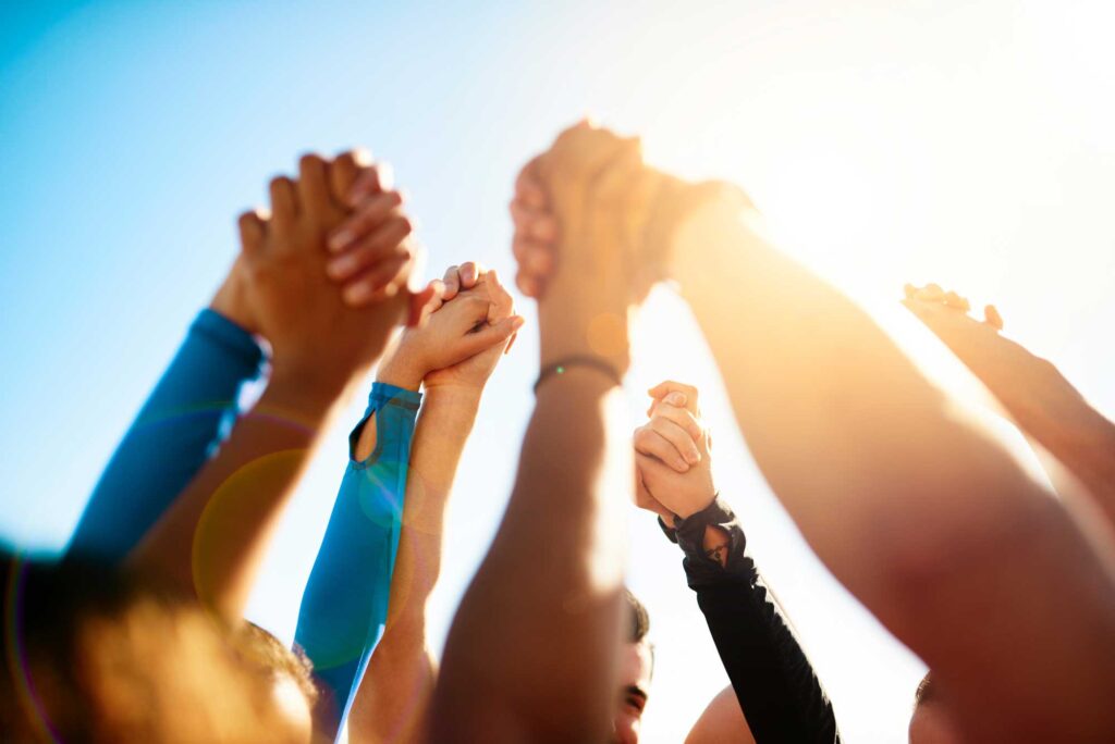 Diverse group of people raising their hands above their heads and clasping hands in celebration, with a bright blue sky and sunshine in the background.