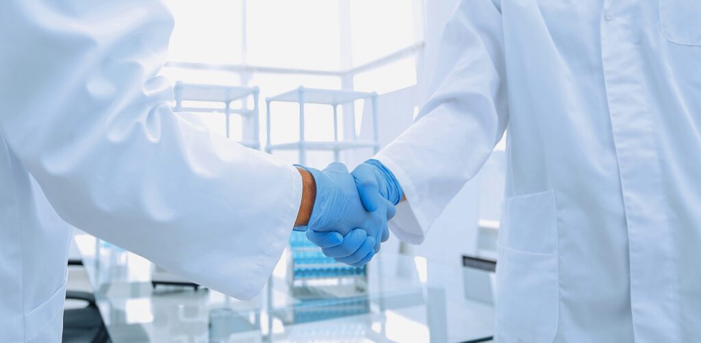 Two scientists wearing lab coats and blue gloves shaking hands in a bright laboratory.