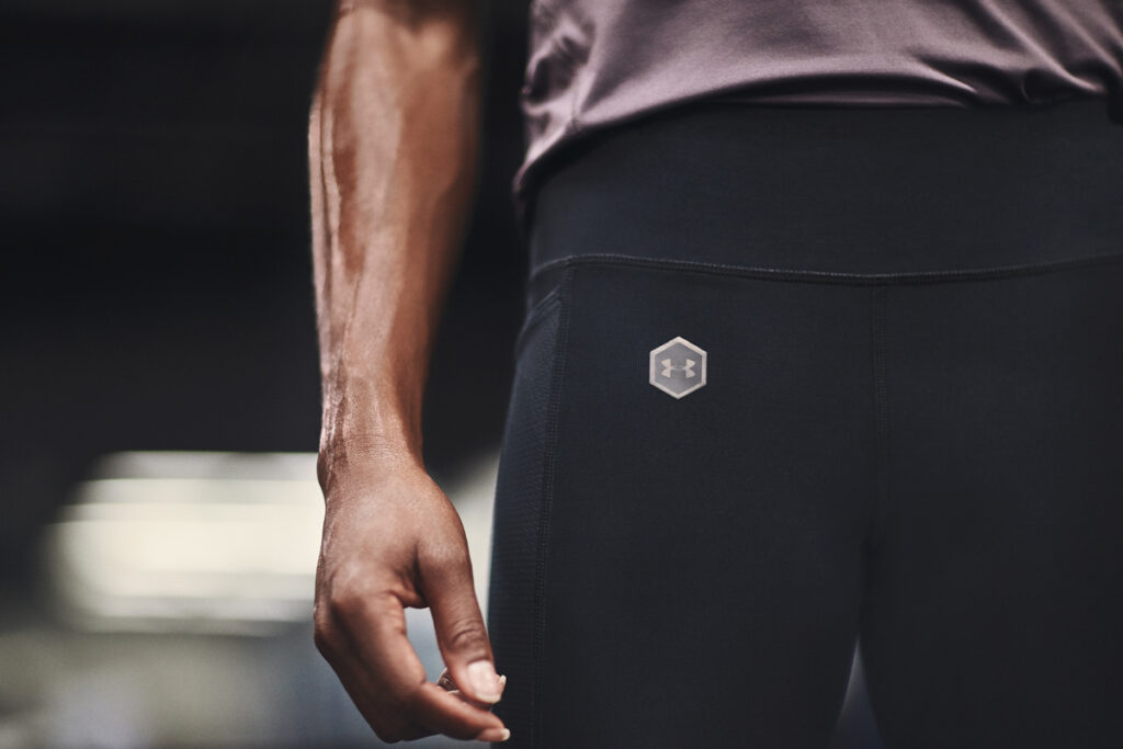 A closeup of the hip of a very fit person wearing Under Armour leggings with logo prominently displayed. Their forearm in the frame is muscular and sweaty.