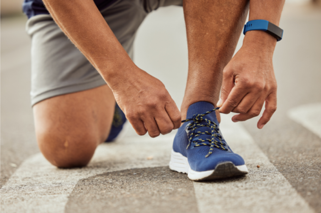 A man kneels down to tie his shoe in a crosswalk. He is wearing activewear and the image highlights his matching blue running shoes and electronic fitness tracker bracelet.