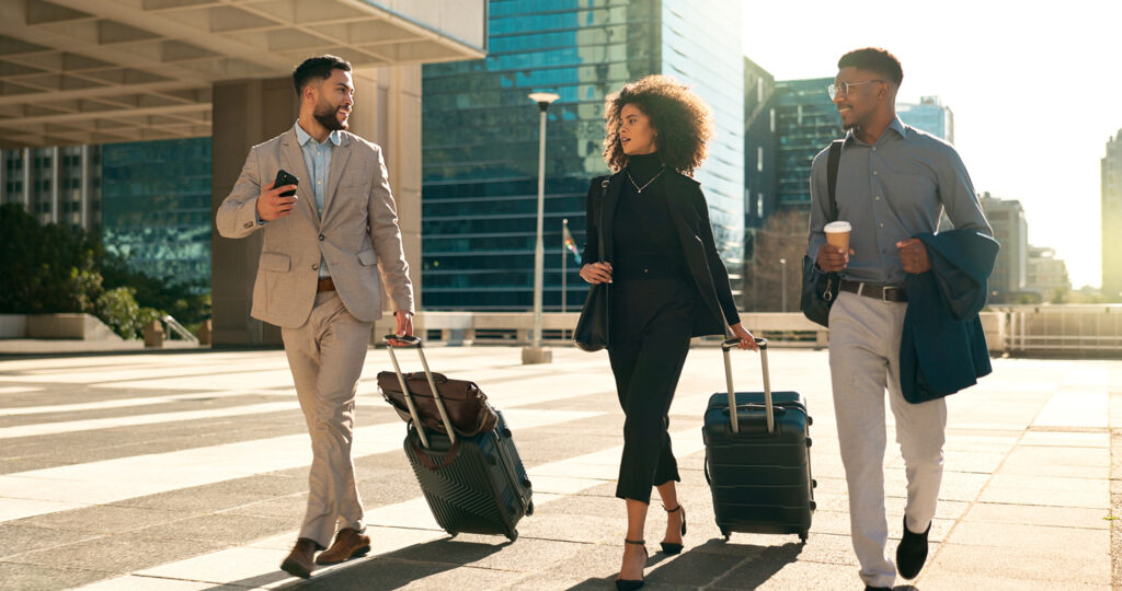 Three professionals walk across a city street wearing fashionable business casual outfits and pulling rolling suitcases behind them.