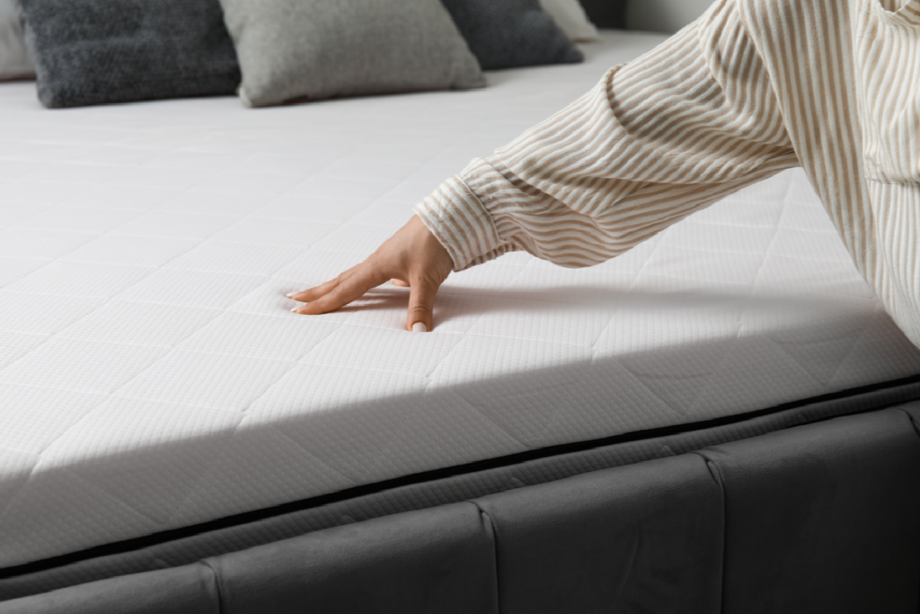 Hand in a striped beige sleeve pressing down on a white mattress surface to test firmness, with gray decorative pillows visible in the background.