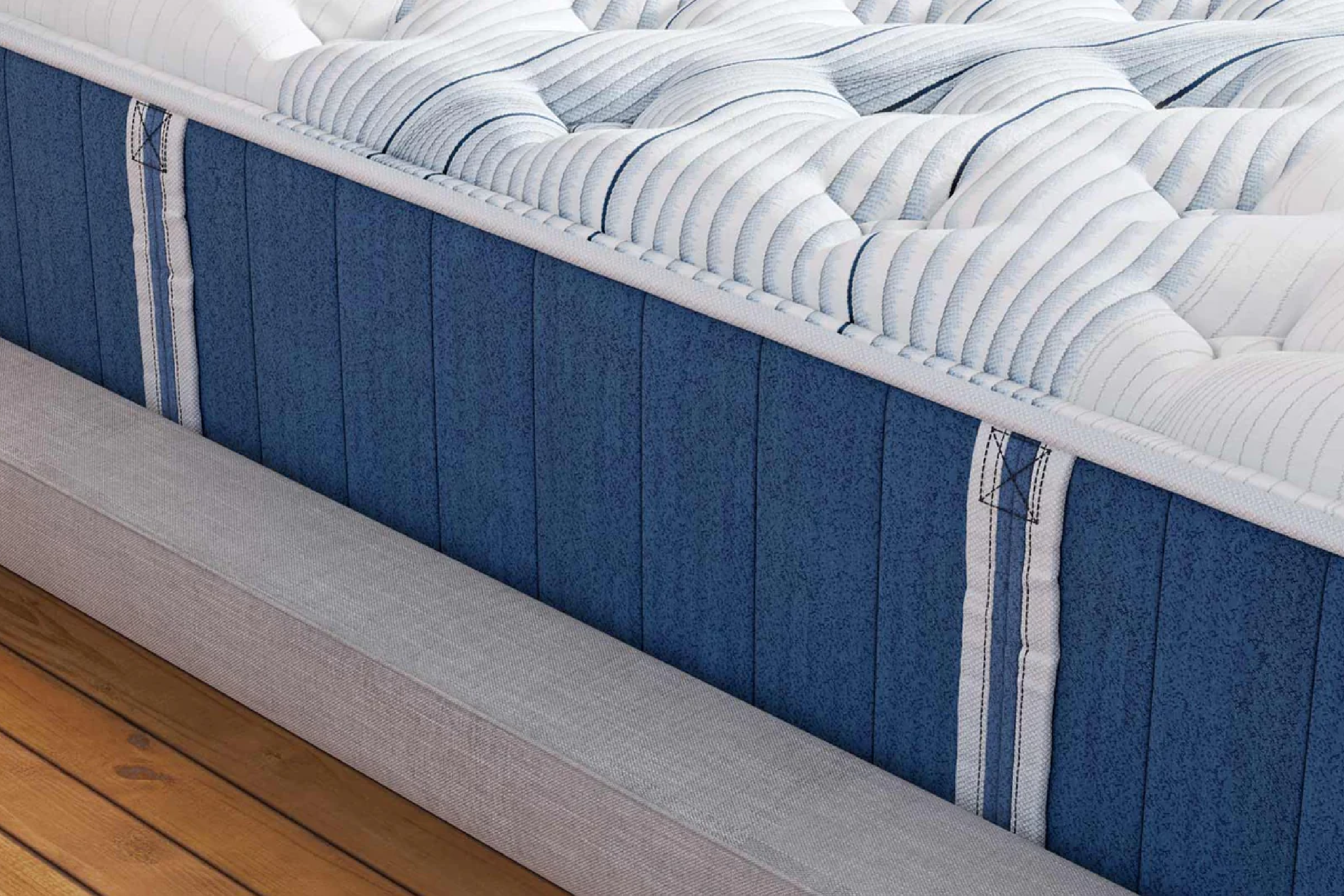 Close-up of a mattress showing a quilted white top layer with ribbed pattern, navy blue side panels, and gray fabric base, sitting on a wooden floor.