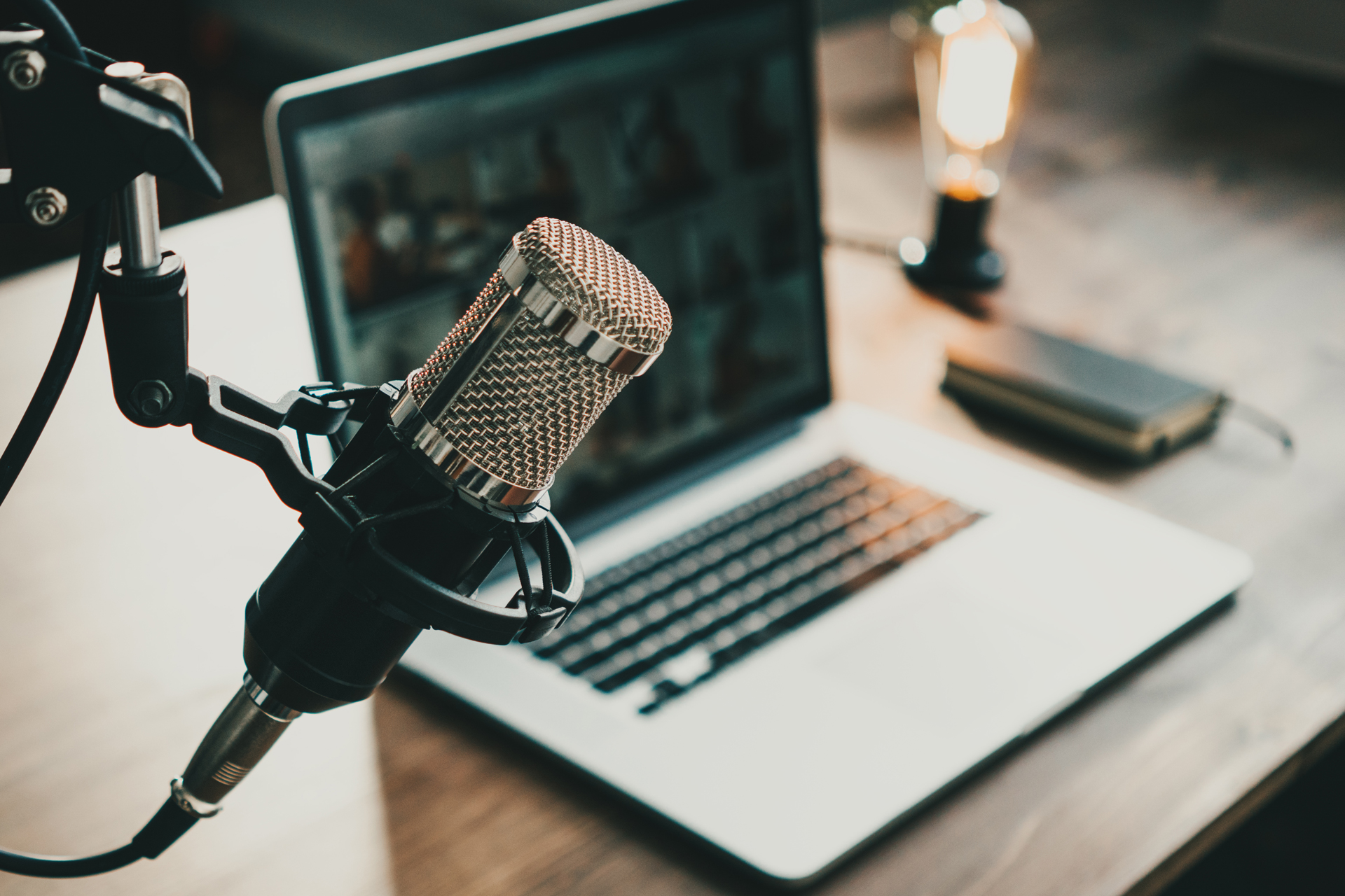 Close-up of a podcast microphone on a stand, with a laptop, notebook, and vintage-style bulb on a desk in the background.