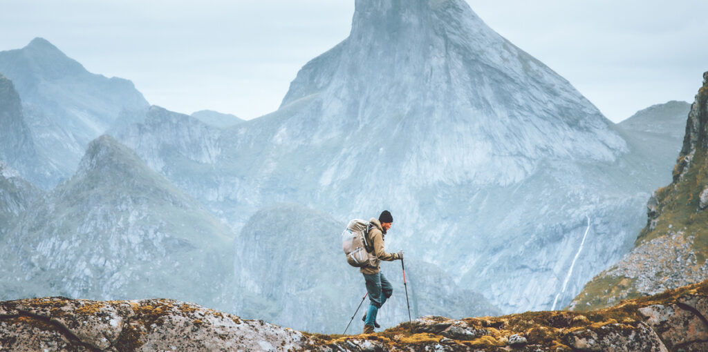 A hiker with trekking poles and a backpack traverses a rocky mountain ridge against a backdrop of towering, misty granite peaks.