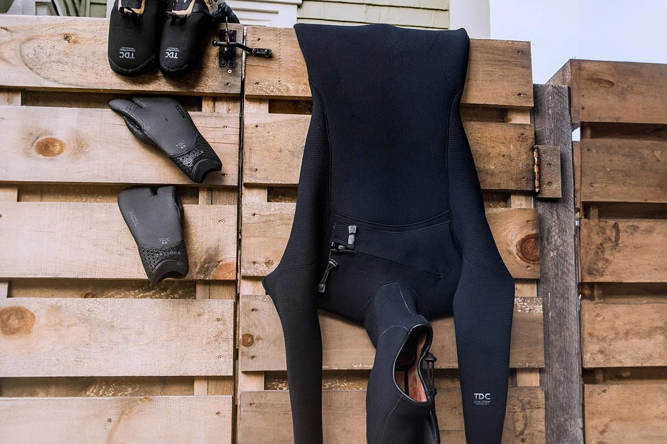 A close-up view of a black TDC wetsuit and gloves hanging on a wooden fence to dry.