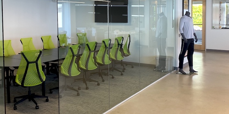 Modern office space with a glass-walled conference room, green ergonomic chairs, and a mannequin wearing athletic apparel.