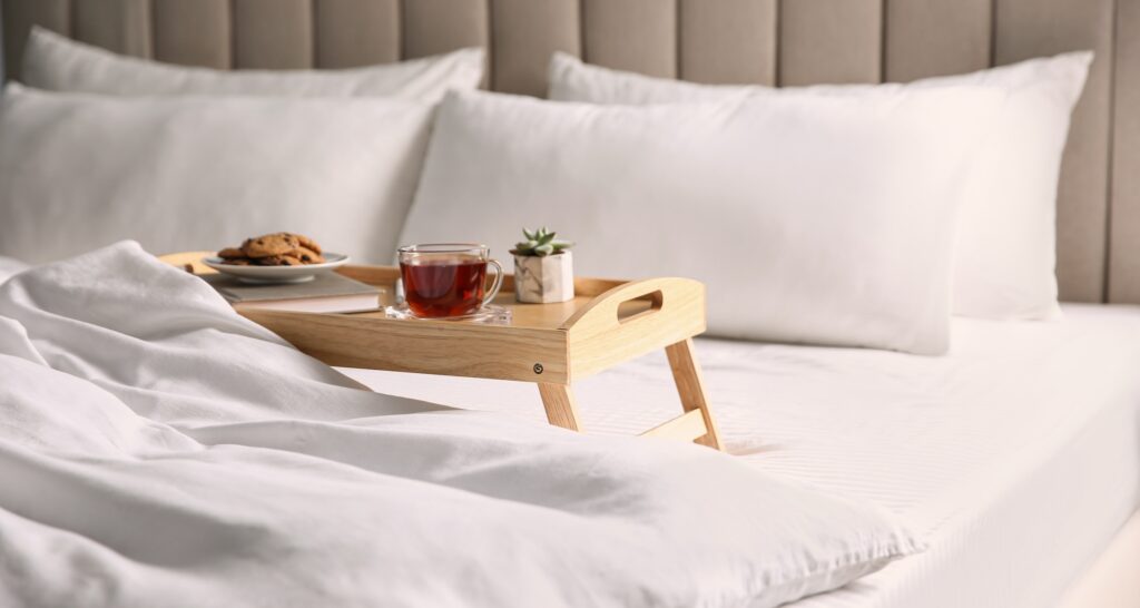 A breakfast-in-bed scene with a wooden tray holding tea, cookies, and a small plant on a white, cozy bed.